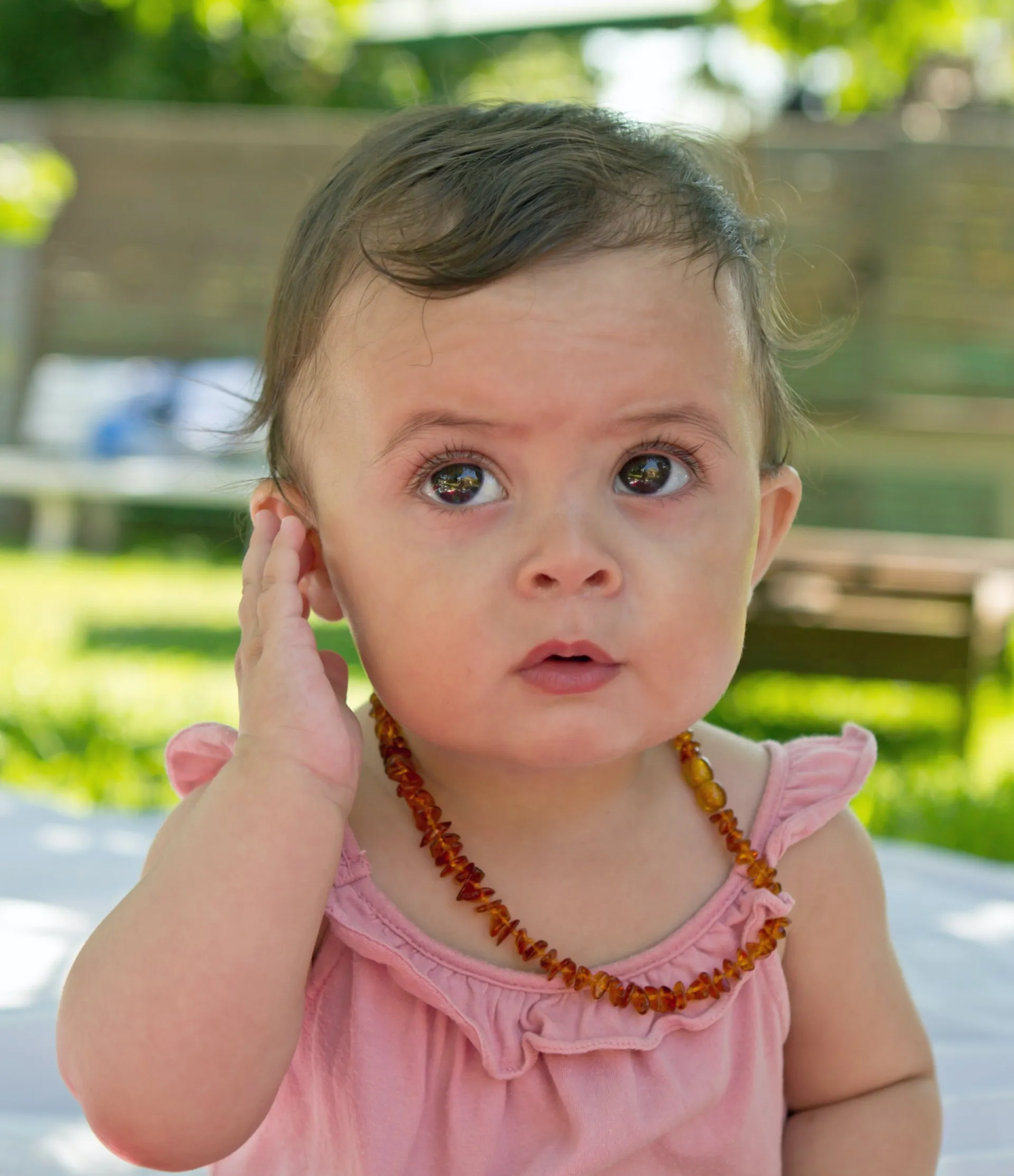 A baby girl wearing an orange necklace and holding her ear.